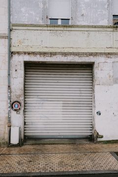 A large weathered metal roller door set in a rough, aged building facade. The structure shows signs of wear with peeling paint and erosion. A small circular sign is mounted to the left of the door, and the area is surrounded by a paved walkway.
