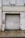 A large weathered metal roller door set in a rough, aged building facade. The structure shows signs of wear with peeling paint and erosion. A small circular sign is mounted to the left of the door, and the area is surrounded by a paved walkway.
