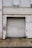 A large weathered metal roller door set in a rough, aged building facade. The structure shows signs of wear with peeling paint and erosion. A small circular sign is mounted to the left of the door, and the area is surrounded by a paved walkway.