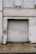 A large weathered metal roller door set in a rough, aged building facade. The structure shows signs of wear with peeling paint and erosion. A small circular sign is mounted to the left of the door, and the area is surrounded by a paved walkway.
