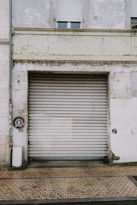 A large weathered metal roller door set in a rough, aged building facade. The structure shows signs of wear with peeling paint and erosion. A small circular sign is mounted to the left of the door, and the area is surrounded by a paved walkway.
