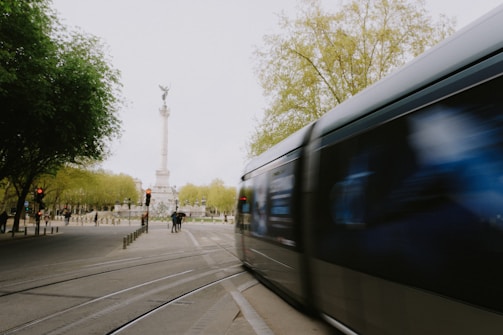 A high-speed train moves along the tracks in an urban park setting. In the background, there is a tall monument with a statue at the top. Trees with fresh green leaves are visible, and a few people stand nearby. The scene captures the motion of the train as it blurs past.