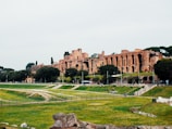 Ancient ruins are set against a backdrop of lush greenery and a cloudy sky. The architecture features large arches and stone structures from a past civilization, surrounded by trees and grass.