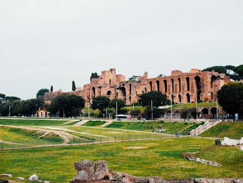 Ancient ruins are set against a backdrop of lush greenery and a cloudy sky. The architecture features large arches and stone structures from a past civilization, surrounded by trees and grass.
