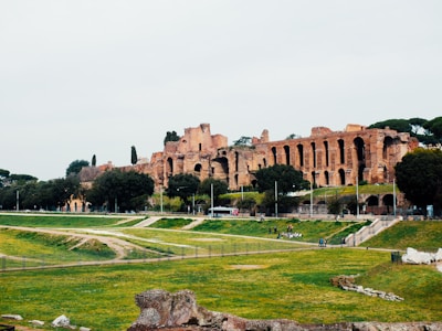 Ancient ruins are set against a backdrop of lush greenery and a cloudy sky. The architecture features large arches and stone structures from a past civilization, surrounded by trees and grass.