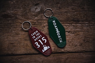 Two retro-style keychains with inscriptions are placed on a rustic wooden surface. The left keychain is maroon and reads 'CABIN IN THE WOODS 315' with a small cabin and tree illustration. The right keychain is green with the text 'ROAMNORTH' printed on it.