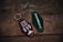 Two retro-style keychains with inscriptions are placed on a rustic wooden surface. The left keychain is maroon and reads 'CABIN IN THE WOODS 315' with a small cabin and tree illustration. The right keychain is green with the text 'ROAMNORTH' printed on it.