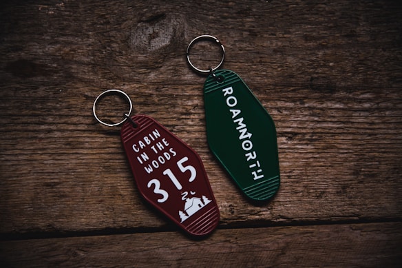 Two retro-style keychains with inscriptions are placed on a rustic wooden surface. The left keychain is maroon and reads 'CABIN IN THE WOODS 315' with a small cabin and tree illustration. The right keychain is green with the text 'ROAMNORTH' printed on it.
