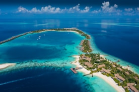 Aerial view of a tropical island surrounded by vibrant turquoise and deep blue waters. The island is dotted with lush greenery and several bungalows or villas. Boats are seen docked near the shore and small pathways connect various parts of the island. Wispy clouds are scattered across the bright blue sky.