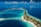 Aerial view of a tropical island surrounded by vibrant turquoise and deep blue waters. The island is dotted with lush greenery and several bungalows or villas. Boats are seen docked near the shore and small pathways connect various parts of the island. Wispy clouds are scattered across the bright blue sky.
