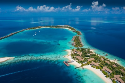 Aerial view of a tropical island surrounded by vibrant turquoise and deep blue waters. The island is dotted with lush greenery and several bungalows or villas. Boats are seen docked near the shore and small pathways connect various parts of the island. Wispy clouds are scattered across the bright blue sky.