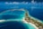 Aerial view of a tropical island surrounded by vibrant turquoise and deep blue waters. The island is dotted with lush greenery and several bungalows or villas. Boats are seen docked near the shore and small pathways connect various parts of the island. Wispy clouds are scattered across the bright blue sky.