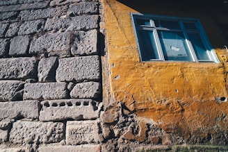 The image captures a close-up view of a building wall, showcasing two distinct textures and colors. One section is constructed with roughly hewn gray stones, while the adjacent part is covered in a yellowish-orange stucco with visible cracks and wear. A window is set into the stucco section, framed in blue, with a curtain visible inside.