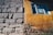 The image captures a close-up view of a building wall, showcasing two distinct textures and colors. One section is constructed with roughly hewn gray stones, while the adjacent part is covered in a yellowish-orange stucco with visible cracks and wear. A window is set into the stucco section, framed in blue, with a curtain visible inside.