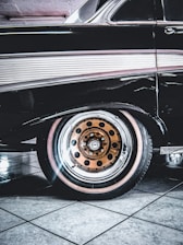 A classic car with a sleek black exterior is highlighted, showcasing its distinctive vintage design. The focus is on the rear wheel, which features a unique orange rim with black circle details. The tile flooring beneath the car is reflective and provides a clean, polished background.