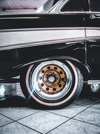 A classic car with a sleek black exterior is highlighted, showcasing its distinctive vintage design. The focus is on the rear wheel, which features a unique orange rim with black circle details. The tile flooring beneath the car is reflective and provides a clean, polished background.