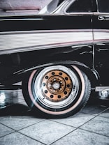 A classic car with a sleek black exterior is highlighted, showcasing its distinctive vintage design. The focus is on the rear wheel, which features a unique orange rim with black circle details. The tile flooring beneath the car is reflective and provides a clean, polished background.