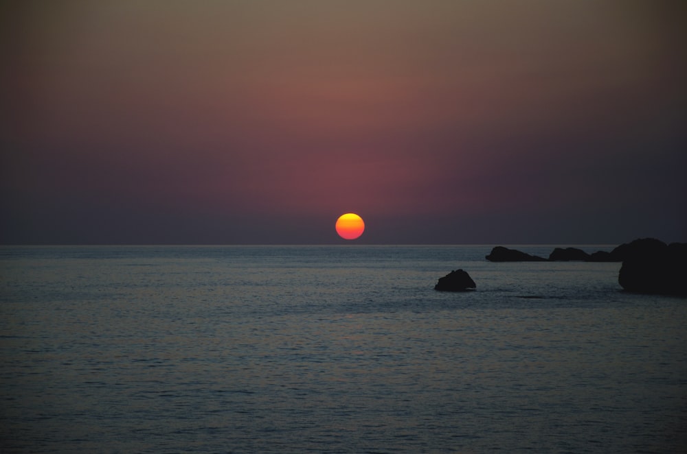 the sun is setting over the ocean with rocks in the foreground