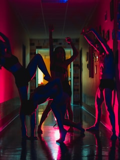 A group of people is performing dynamic dance poses in a dimly lit hallway, with silhouettes enhanced by vibrant red and blue lighting. The poses are expressive and artistic, creating a visually striking scene.
