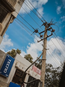 An electrical pole with multiple wires and insulators extends upwards against a partly cloudy sky. The building on the left has signage for 'Tecno' and 'Mi' brands. Trees are visible in the background.