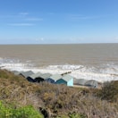 Several small beach huts with sloped roofs sit on a grassy hillside overlooking a vast expanse of ocean. The sea is dotted with white waves, and the horizon is visible under a clear blue sky.