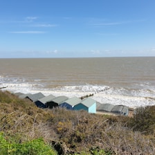 Several small beach huts with sloped roofs sit on a grassy hillside overlooking a vast expanse of ocean. The sea is dotted with white waves, and the horizon is visible under a clear blue sky.