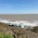 Several small beach huts with sloped roofs sit on a grassy hillside overlooking a vast expanse of ocean. The sea is dotted with white waves, and the horizon is visible under a clear blue sky.