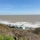 Several small beach huts with sloped roofs sit on a grassy hillside overlooking a vast expanse of ocean. The sea is dotted with white waves, and the horizon is visible under a clear blue sky.