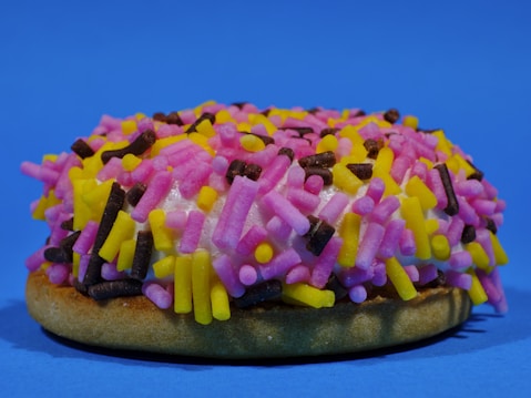 A frosted doughnut is topped with an assortment of colorful sprinkles in pink, yellow, and brown shades. The backdrop is a solid bright blue, creating a vibrant contrast with the doughnut's vivid topping.