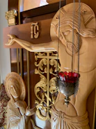 A detailed wooden carving of a crucifix with Jesus Christ, featuring intricate designs and a rich golden color. Next to it, a depiction of a holy figure, possibly Mary, looks upward. A decorative hanging lamp with red glass elements is prominently placed in front.