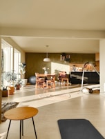 A cozy living space is filled with natural light streaming in from large windows. There are several plants scattered throughout the room, adding a touch of greenery. A wooden dining table with chairs is placed near a wall adorned with shelves and a few decorative items. A pendant lamp hangs above the table. On the floor, a round woven rug and a black exercise mat are visible, alongside a small round coffee table with thin legs. A comfortable atmosphere pervades the room, enhanced by light-colored walls and a warm tone.