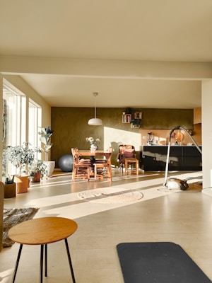 A cozy living space is filled with natural light streaming in from large windows. There are several plants scattered throughout the room, adding a touch of greenery. A wooden dining table with chairs is placed near a wall adorned with shelves and a few decorative items. A pendant lamp hangs above the table. On the floor, a round woven rug and a black exercise mat are visible, alongside a small round coffee table with thin legs. A comfortable atmosphere pervades the room, enhanced by light-colored walls and a warm tone.