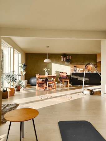 A cozy living space is filled with natural light streaming in from large windows. There are several plants scattered throughout the room, adding a touch of greenery. A wooden dining table with chairs is placed near a wall adorned with shelves and a few decorative items. A pendant lamp hangs above the table. On the floor, a round woven rug and a black exercise mat are visible, alongside a small round coffee table with thin legs. A comfortable atmosphere pervades the room, enhanced by light-colored walls and a warm tone.
