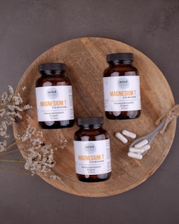 Three bottles of magnesium supplements are placed on a rustic wooden platter along with a small spoon containing white capsules. Dried flowers are arranged artfully around the bottles. The background is a textured, dark gray surface.