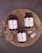 Three bottles of magnesium supplements are placed on a rustic wooden platter along with a small spoon containing white capsules. Dried flowers are arranged artfully around the bottles. The background is a textured, dark gray surface.