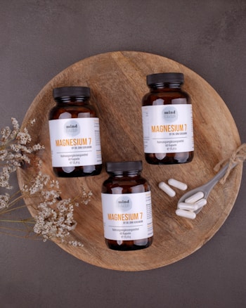 Three bottles of magnesium supplements are placed on a rustic wooden platter along with a small spoon containing white capsules. Dried flowers are arranged artfully around the bottles. The background is a textured, dark gray surface.