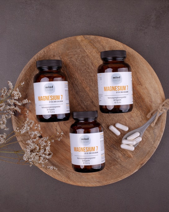 Three bottles of magnesium supplements are placed on a rustic wooden platter along with a small spoon containing white capsules. Dried flowers are arranged artfully around the bottles. The background is a textured, dark gray surface.