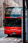 A bright red double-decker tourist bus with 'SKY BUS TOKYO' on the front is parked on the side of an urban street. The bus features multiple circular headlights and a sleek design. Nearby, a bus stop sign is visible, and various buildings with modern architecture line the street in the background.