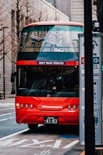 A bright red double-decker tourist bus with 'SKY BUS TOKYO' on the front is parked on the side of an urban street. The bus features multiple circular headlights and a sleek design. Nearby, a bus stop sign is visible, and various buildings with modern architecture line the street in the background.