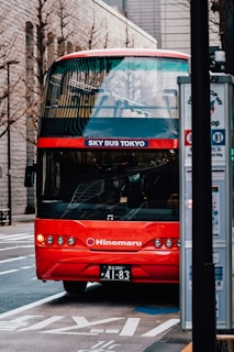 A bright red double-decker tourist bus with 'SKY BUS TOKYO' on the front is parked on the side of an urban street. The bus features multiple circular headlights and a sleek design. Nearby, a bus stop sign is visible, and various buildings with modern architecture line the street in the background.