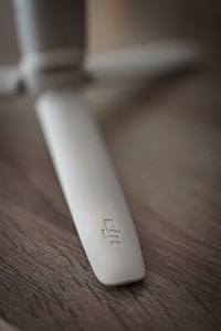 A close-up shot of a light gray plastic propeller with a logo on it, set against a textured wooden surface. The focus is on the propeller blade, which appears sleek and slightly curved.