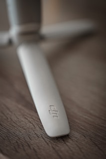 A close-up shot of a light gray plastic propeller with a logo on it, set against a textured wooden surface. The focus is on the propeller blade, which appears sleek and slightly curved.