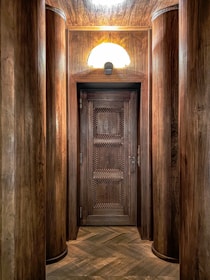 An ornate wooden door with a distinct geometric pattern is framed by large, cylindrical wooden columns. The lighting above casts a warm glow on the wood, highlighting its rich textures and tones. The floor is laid with a herringbone wood pattern that complements the overall wooden theme.