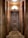 An ornate wooden door with a distinct geometric pattern is framed by large, cylindrical wooden columns. The lighting above casts a warm glow on the wood, highlighting its rich textures and tones. The floor is laid with a herringbone wood pattern that complements the overall wooden theme.