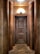 An ornate wooden door with a distinct geometric pattern is framed by large, cylindrical wooden columns. The lighting above casts a warm glow on the wood, highlighting its rich textures and tones. The floor is laid with a herringbone wood pattern that complements the overall wooden theme.