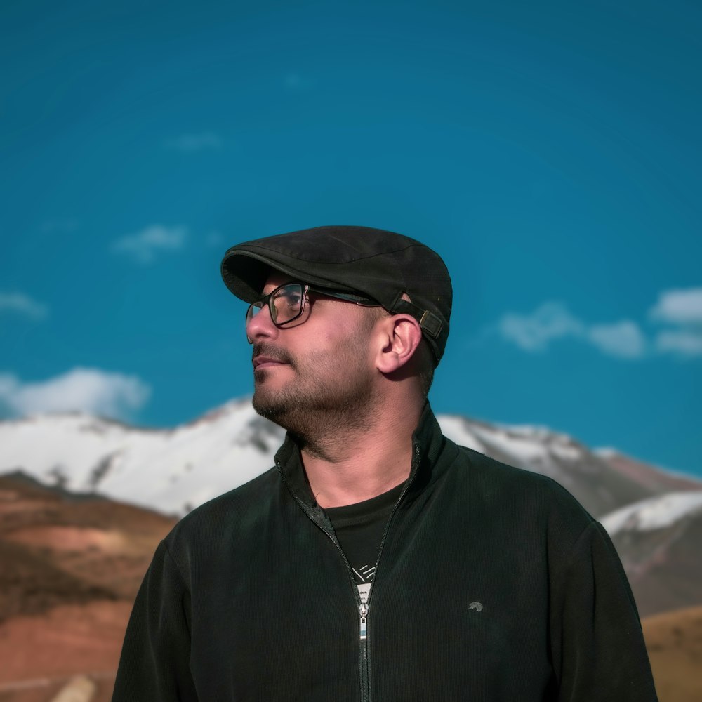 a man wearing a hat and glasses standing in front of a mountain
