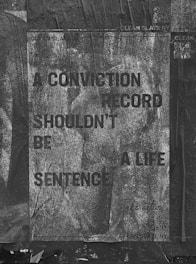 A weathered poster with text that reads 'A CONVICTION RECORD SHOULDN'T BE A LIFE SENTENCE' is displayed. The background is textured with a grayscale, worn look, and the poster appears to be pasted on a rough, dark surface. Some QR codes and text related to 'Clean Slate' are visible.
