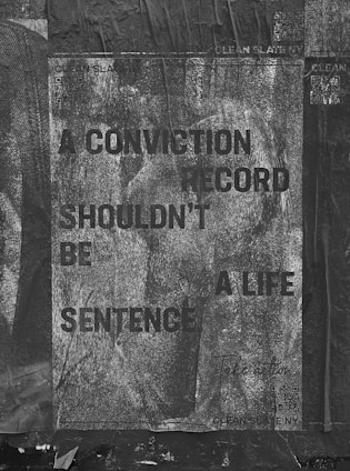 A weathered poster with text that reads 'A CONVICTION RECORD SHOULDN'T BE A LIFE SENTENCE' is displayed. The background is textured with a grayscale, worn look, and the poster appears to be pasted on a rough, dark surface. Some QR codes and text related to 'Clean Slate' are visible.