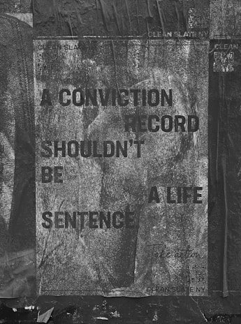 A weathered poster with text that reads 'A CONVICTION RECORD SHOULDN'T BE A LIFE SENTENCE' is displayed. The background is textured with a grayscale, worn look, and the poster appears to be pasted on a rough, dark surface. Some QR codes and text related to 'Clean Slate' are visible.