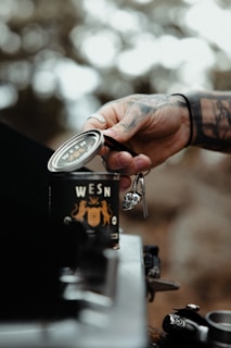 A tattooed hand is holding a can labeled with the word 'WESN', set against a blurred natural background. The scene has a rustic feel with blurred trees in the background, suggesting an outdoor setting.